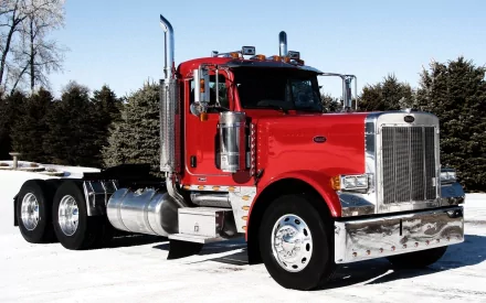 HD desktop wallpaper featuring a bright red Peterbilt truck parked on snow with trees in the background, showcasing the vehicle's classic design and powerful presence.