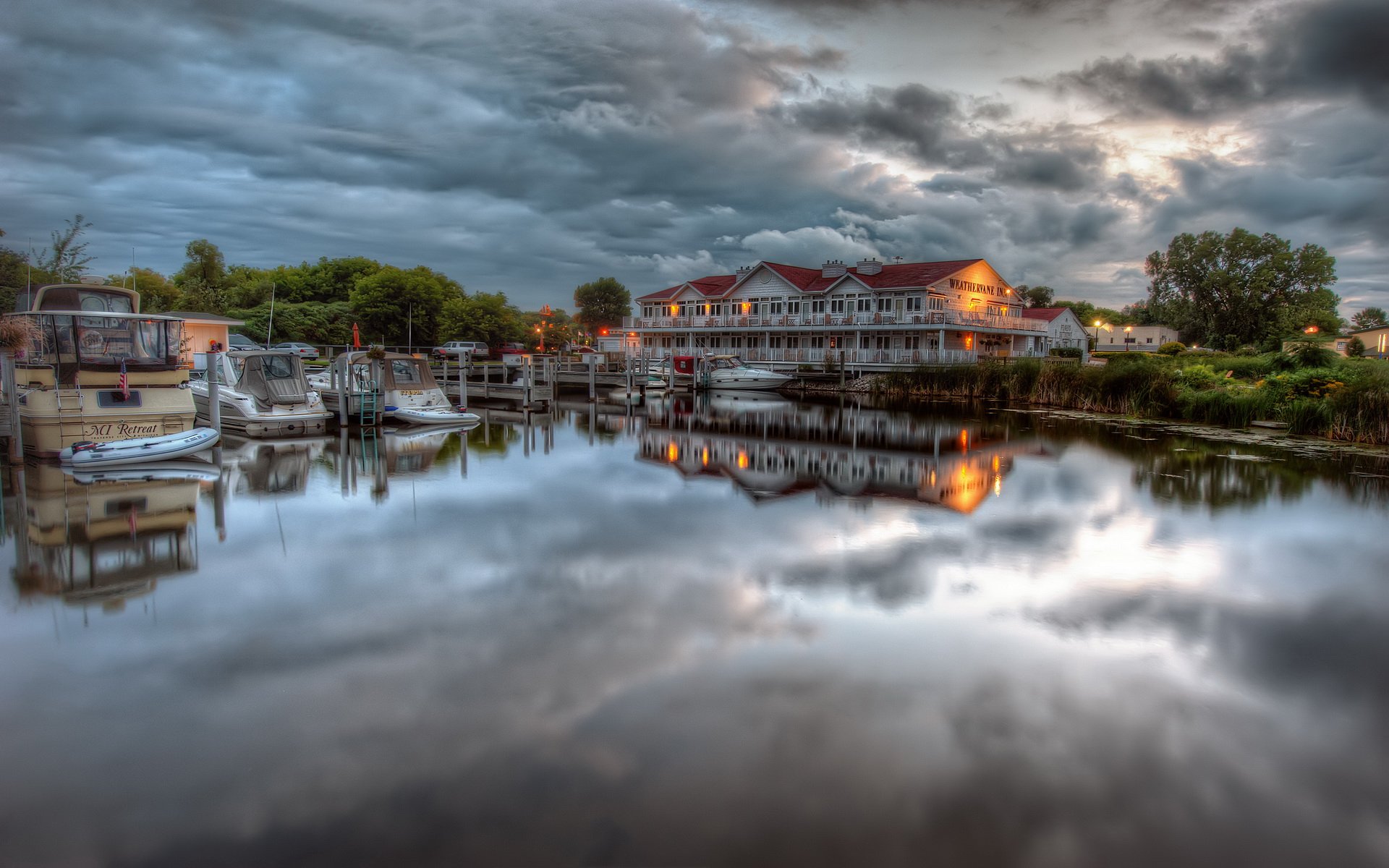 HD desktop wallpaper photograph of a waterfront marina at dusk with moored boats and a lit boathouse reflected in calm water beneath a dramatic clouded sky.