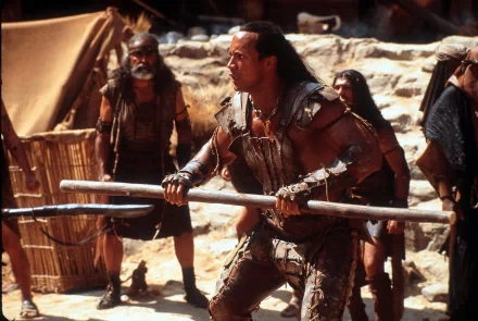Muscular desert warrior in leather armor grips twin staves amid a sandbound camp, cinematic still from the movie The Scorpion King — 2K Quad HD PC desktop wallpaper and background.