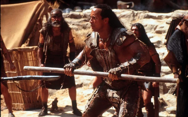 Muscular desert warrior in leather armor grips twin staves amid a sandbound camp, cinematic still from the movie The Scorpion King — 2K Quad HD PC desktop wallpaper and background.