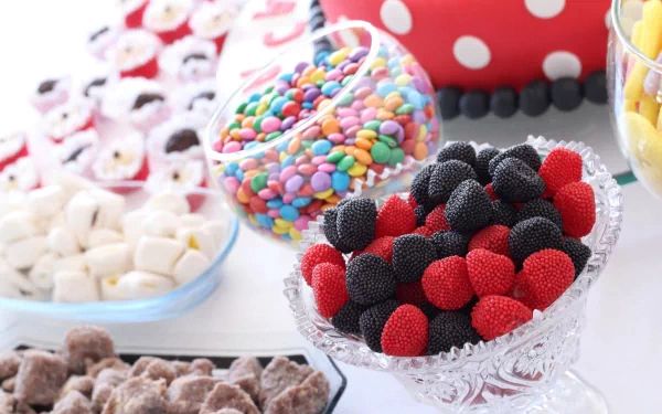 A colorful assortment of candies displayed in various bowls, featuring red and black gummy candies, vibrant chocolate-coated candies, and fluffy marshmallows, set against a cheerful background.