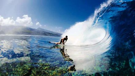 A surfer skillfully rides a large wave on a clear, sunny day, with mountains in the background and clear blue water below. This high-definition image makes an engaging desktop wallpaper.