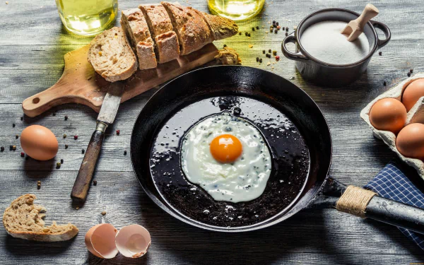 A close-up of a fried egg in a black skillet, surrounded by fresh eggs, slices of bread, a bowl of salt, and cooking utensils, set against a rustic wooden background. 4K Ultra HD wallpaper.