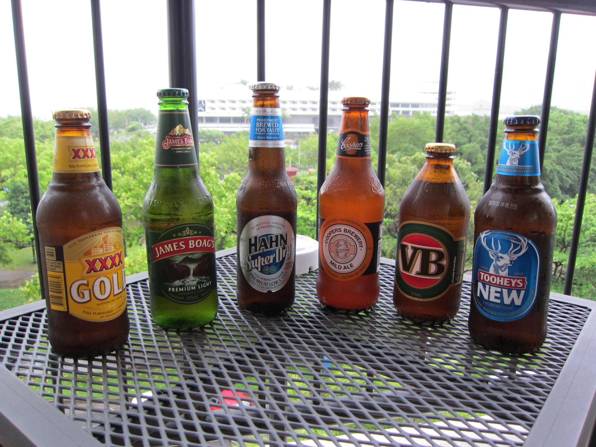 HD desktop wallpaper featuring six diverse beer bottles displayed on a black metal table outdoors with lush greenery in the background.