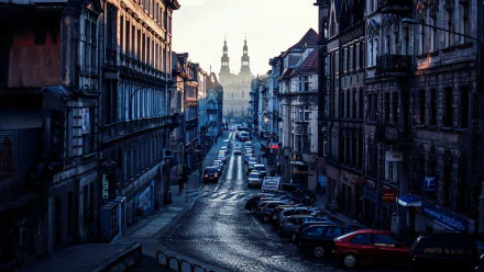 A scenic view of a cobblestone street in Poznań, lined with historic buildings and parked cars, leading towards a distant silhouette of twin towers under a soft morning light.