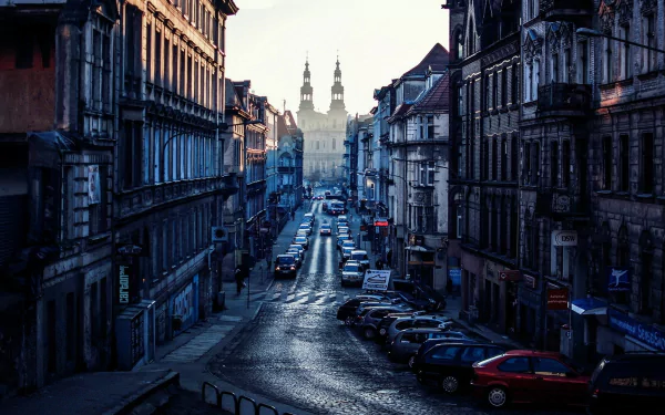 A scenic view of a cobblestone street in Poznań, lined with historic buildings and parked cars, leading towards a distant silhouette of twin towers under a soft morning light.