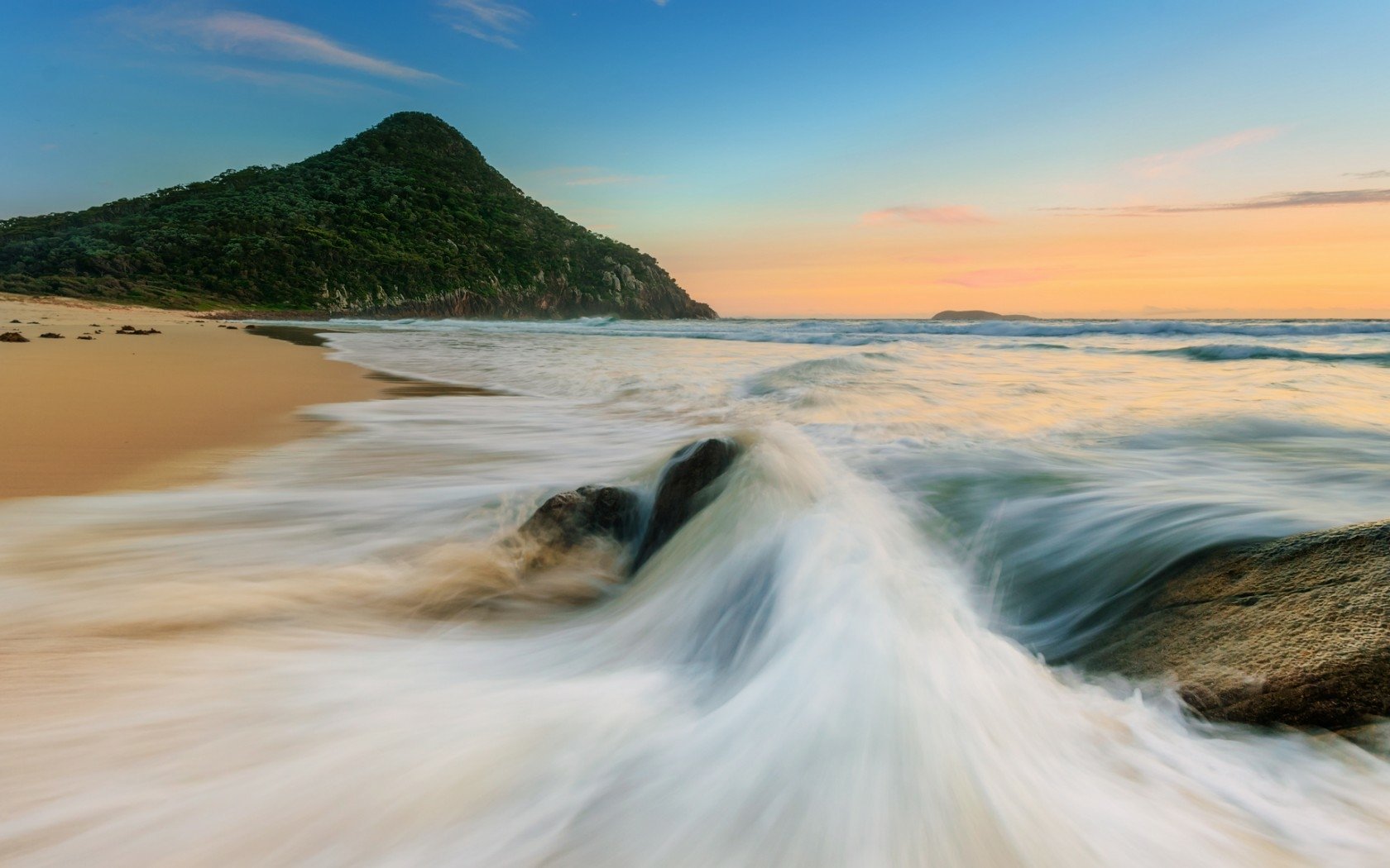 HD PC desktop wallpaper and background: nature beach at sunset, waves rushing over rocks to a sandy shore with a green hill on the horizon.