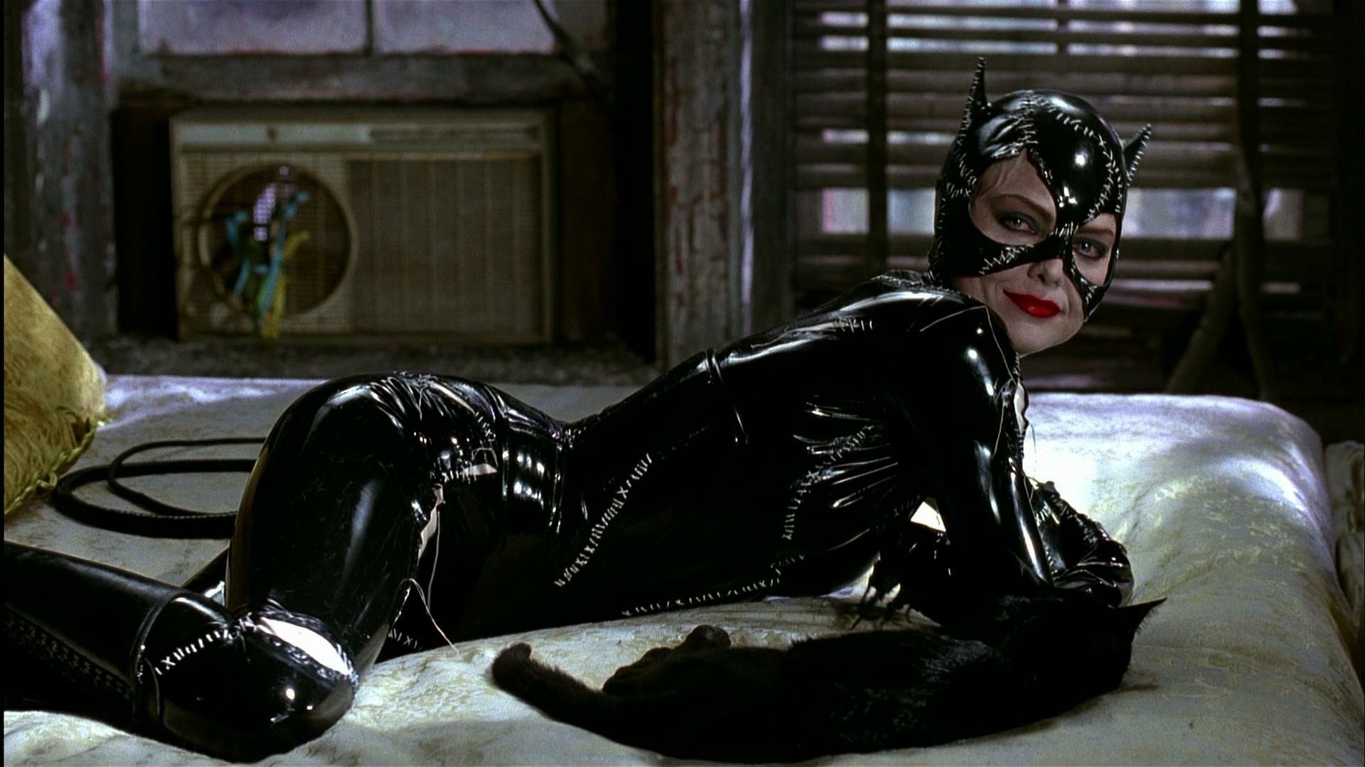 HD PC desktop wallpaper featuring Catwoman in her black latex suit from the movie Batman Returns, lying on a bed in a dimly lit room.