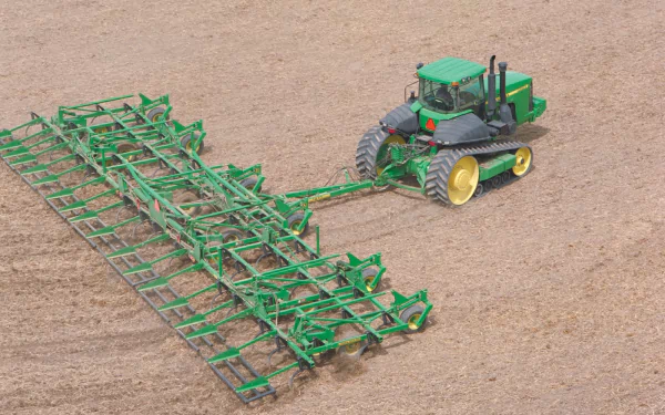 HD desktop wallpaper featuring a John Deere vehicle towing large green farming equipment across a plowed field.