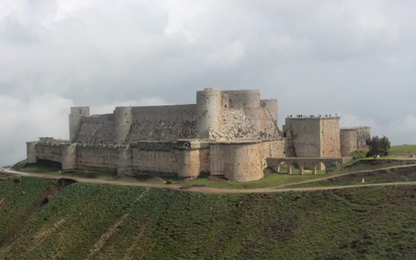 HD PC wallpaper featuring Krak des Chevaliers, a massive man-made medieval fortress standing on a lush green hill under a cloudy sky.