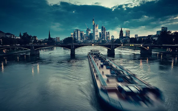 HD desktop wallpaper of Frankfurt's skyline at dusk, featuring the river, a bridge, and a blurred boat in motion, showcasing man-made urban architecture.