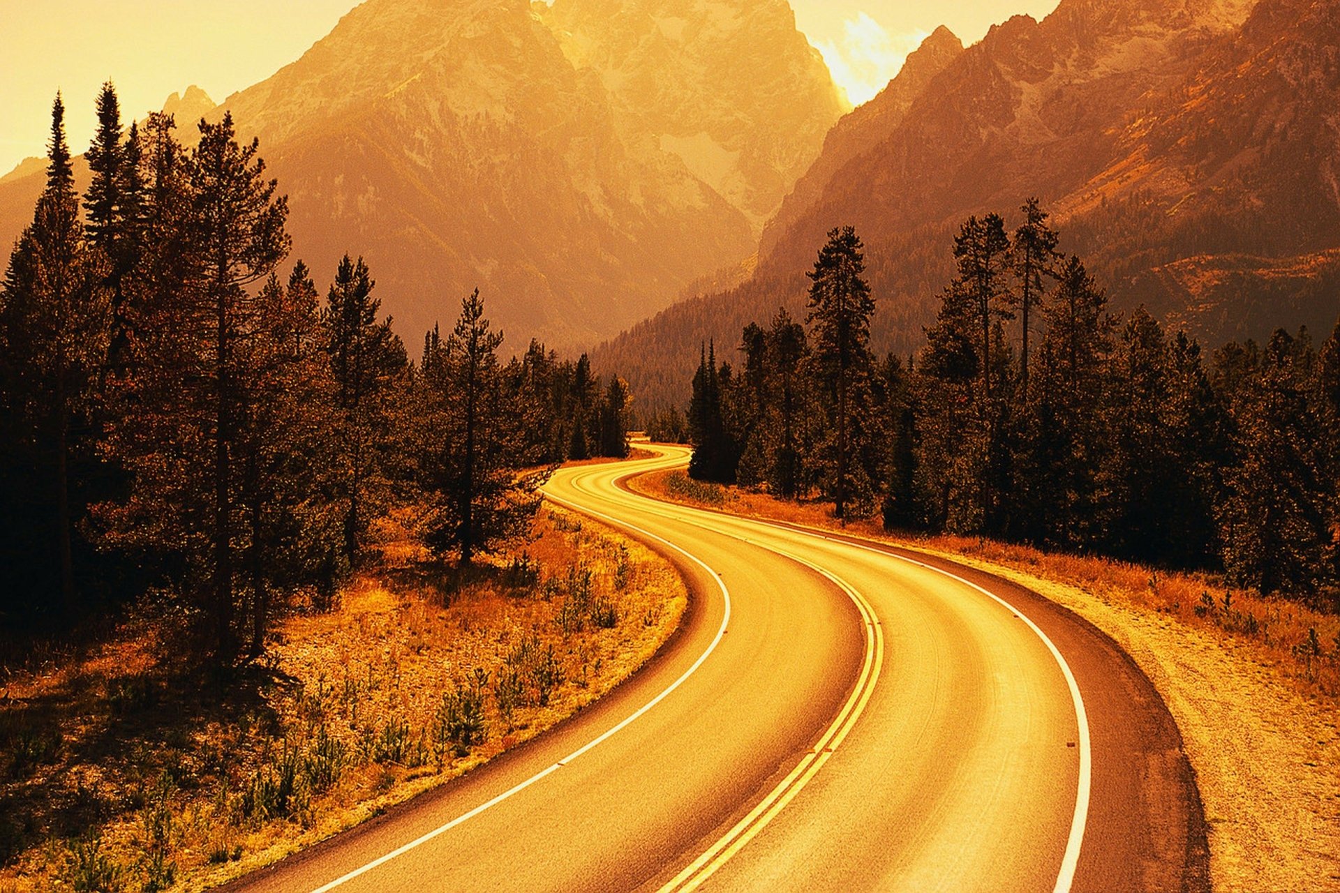 HD PC desktop wallpaper showing a winding man-made road through a forested area with mountains in the background bathed in warm golden light.
