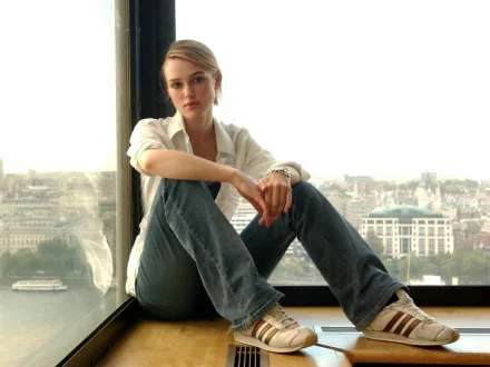 A serene HD desktop wallpaper featuring celebrity Keira Knightley sitting by a large window, overlooking a city skyline, dressed casually in jeans and sneakers.