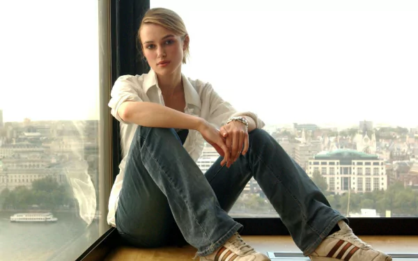 A serene HD desktop wallpaper featuring celebrity Keira Knightley sitting by a large window, overlooking a city skyline, dressed casually in jeans and sneakers.