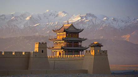 HD wallpaper showing the man-made Jiayu Pass fortress with traditional Chinese architecture set against a backdrop of snow-capped mountains during sunset.