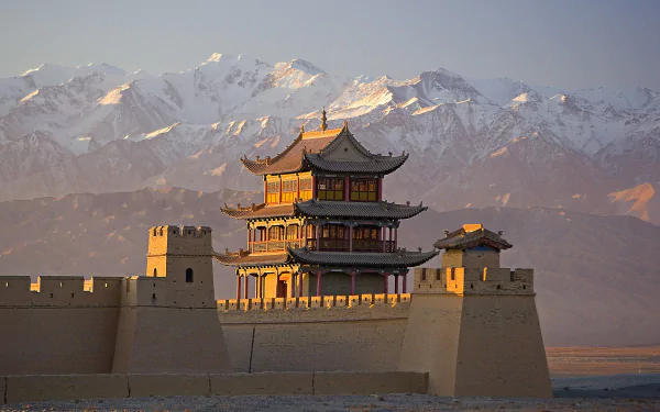 HD wallpaper showing the man-made Jiayu Pass fortress with traditional Chinese architecture set against a backdrop of snow-capped mountains during sunset.