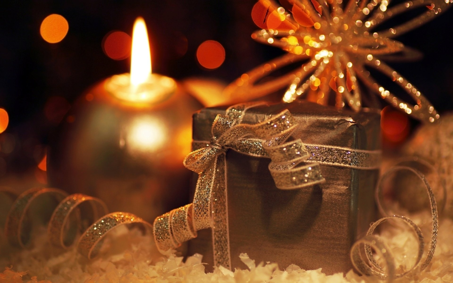 A warm, festive scene featuring a lit candle alongside a beautifully wrapped Christmas gift, adorned with a glittery ribbon, set against a backdrop of holiday lights.