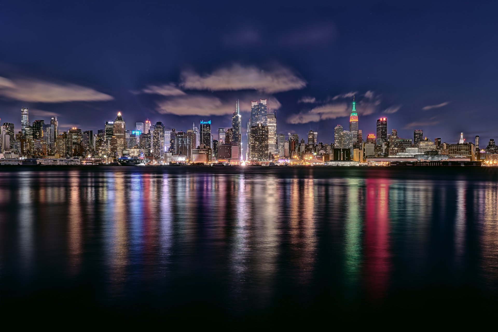 HD PC desktop wallpaper showcasing the illuminated New York City skyline at night, reflecting vibrant city lights on the water under a partly cloudy sky.