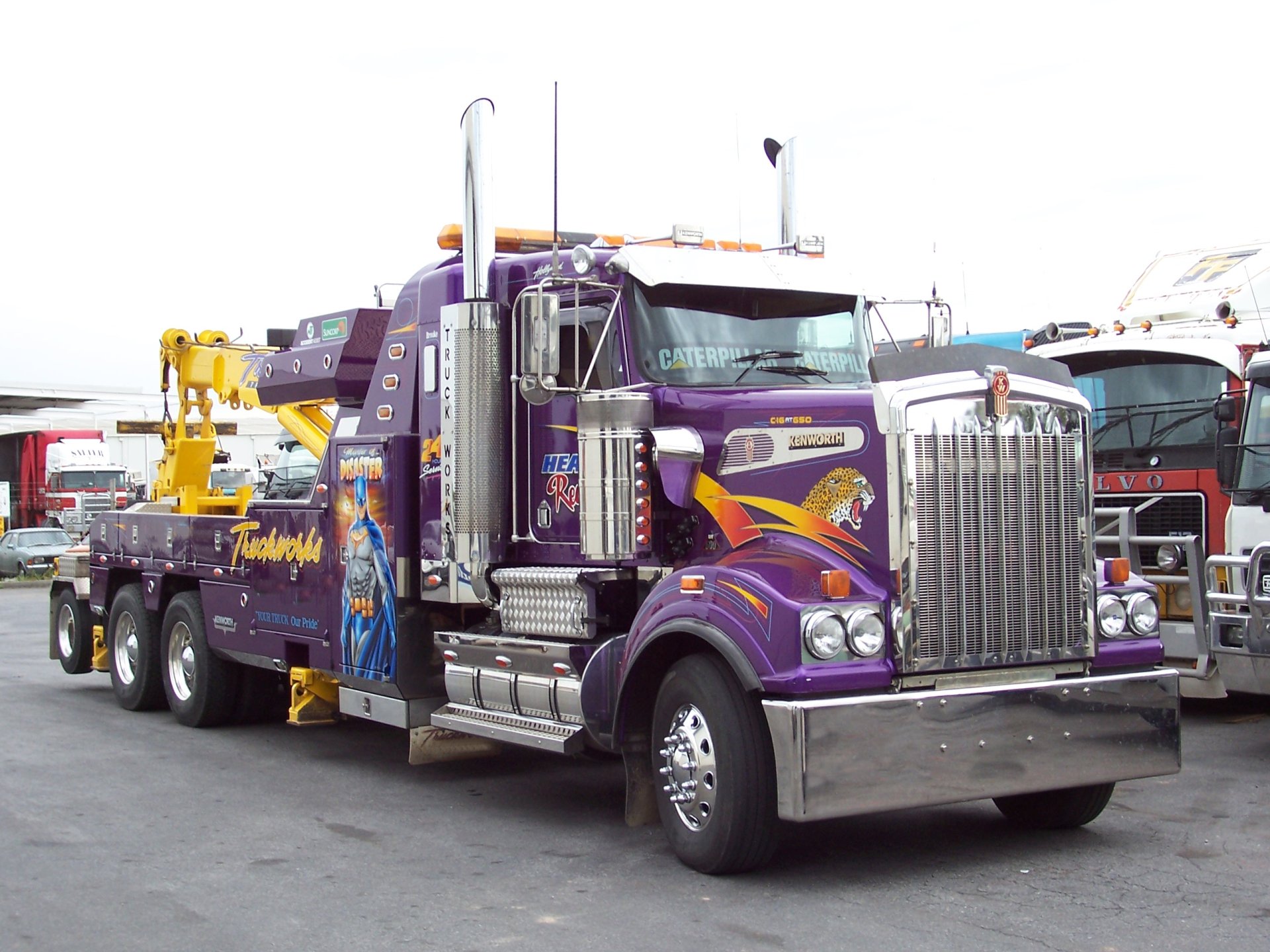 HD desktop wallpaper featuring a purple Kenworth tow truck with detailed graphics, parked among other trucks in a lot.