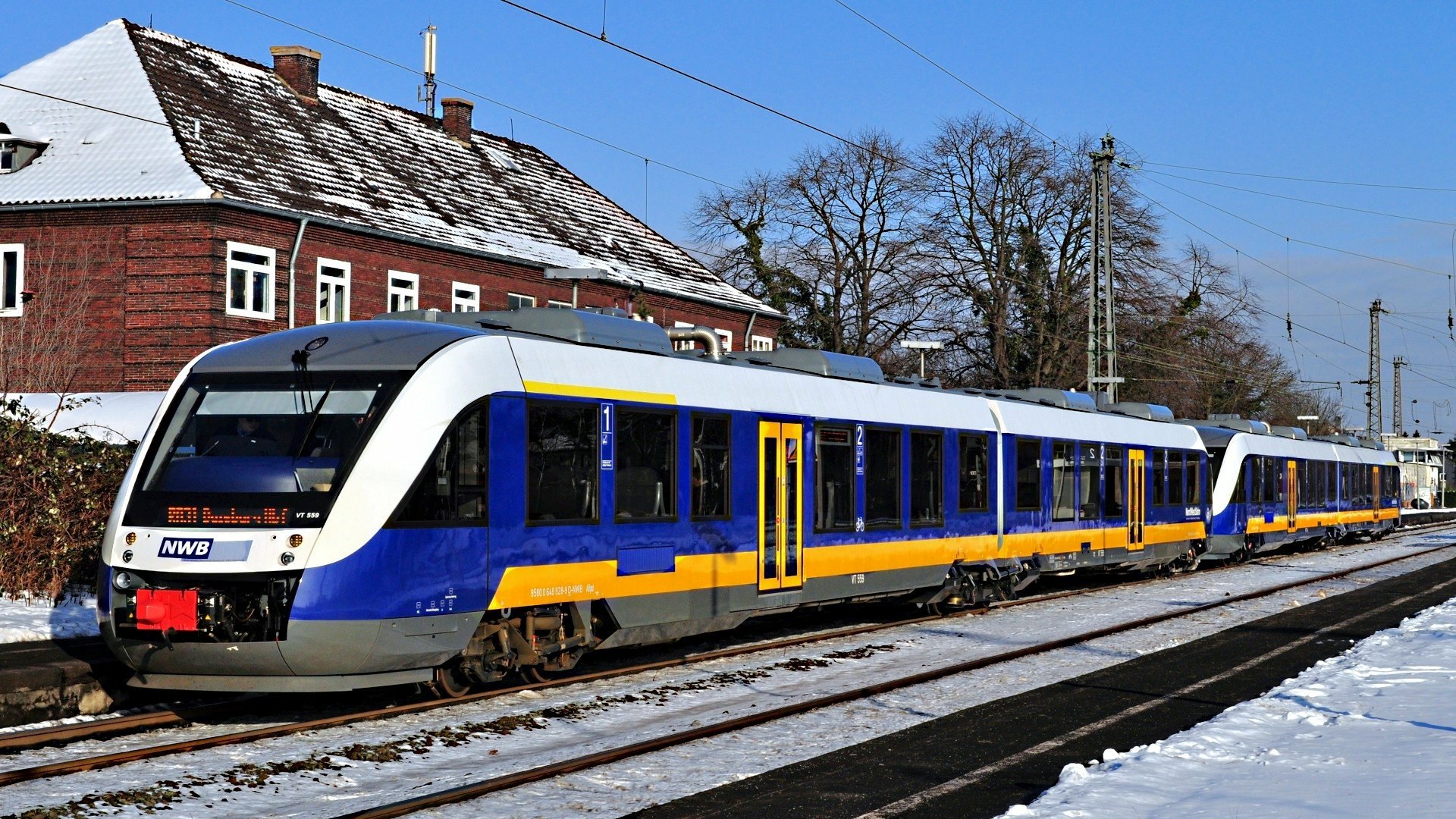 A sleek, modern train in blue and yellow colors sits on snowy tracks, surrounded by trees and buildings. This vibrant image serves as an HD wallpaper for train enthusiasts.