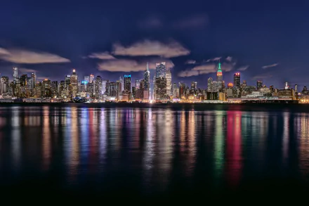 HD PC desktop wallpaper showcasing the illuminated New York City skyline at night, reflecting vibrant city lights on the water under a partly cloudy sky.