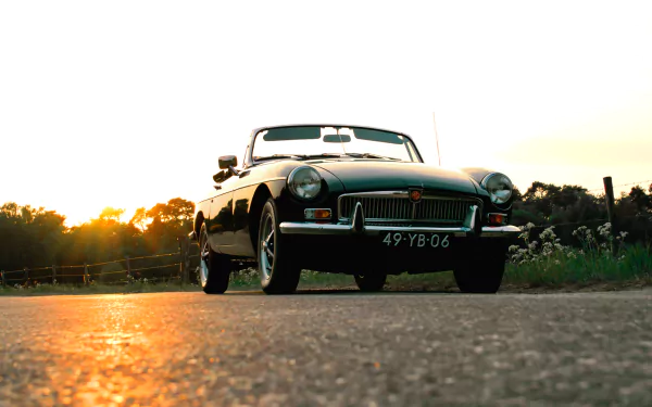 HD PC desktop wallpaper featuring a classic MG vehicle captured low to the ground with a sunset backdrop on a rural road.