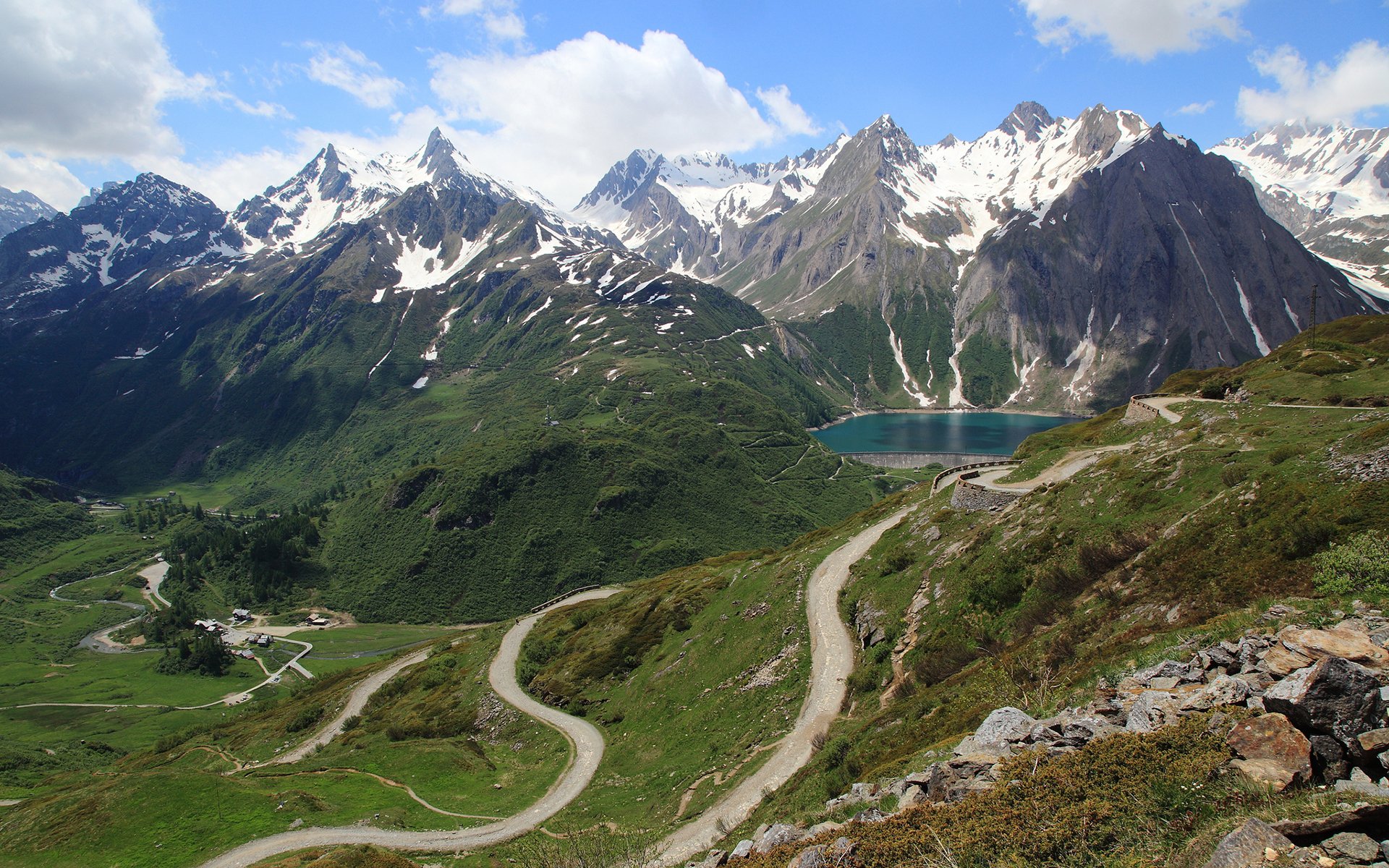 HD desktop wallpaper featuring winding mountain roads, lush green valleys, snow-capped peaks, and a serene alpine lake under a bright blue sky.