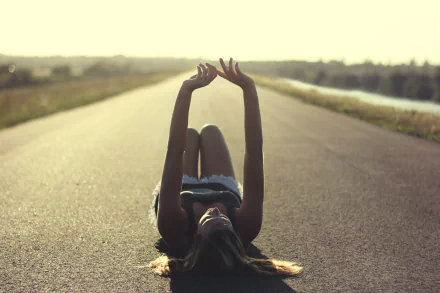 HD desktop wallpaper of a woman lying on a road, stretching her arms up, capturing a serene mood in a sunlit rural setting.