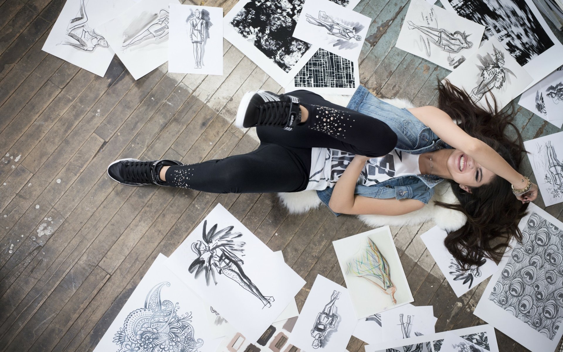 A stylish HD wallpaper featuring Selena Gomez lying among sketches and art on a wooden floor, showcasing her playful and artistic side in a vibrant setting.