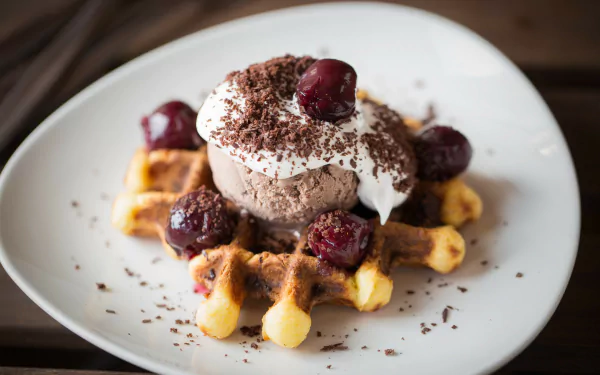HD desktop wallpaper showing a close-up of a waffle topped with chocolate ice cream, whipped cream, cherries, and chocolate shavings on a white plate.