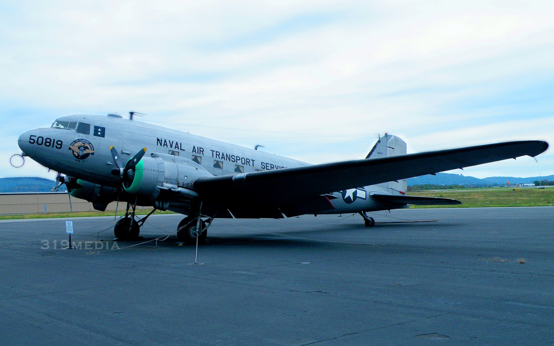 HD desktop wallpaper featuring a military Douglas DC-3 aircraft parked on an airfield under a cloudy sky, showcasing vintage naval air transport photography.