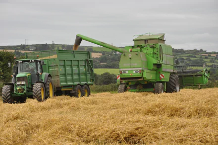 HD desktop wallpaper featuring John Deere agricultural vehicles harvesting a golden field under an overcast sky.