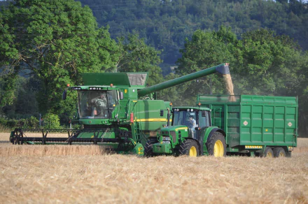 HD PC desktop wallpaper showing a John Deere combine harvester unloading grain into a tractor trailer in a golden field with green trees and hills in the background.