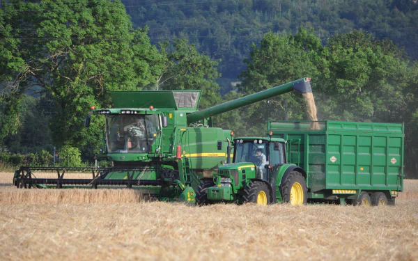 HD PC desktop wallpaper showing a John Deere combine harvester unloading grain into a tractor trailer in a golden field with green trees and hills in the background.