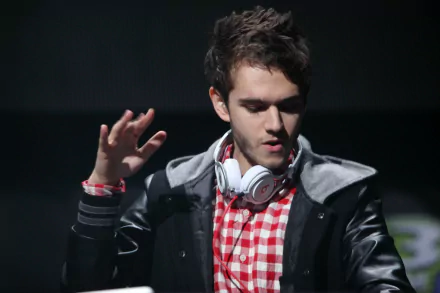 HD PC wallpaper of a music-themed DJ performing on stage: young man in a red checkered shirt and headphones around his neck, raising a hand against a dark background.