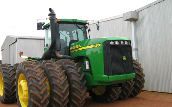 A John Deere tractor with large tires is positioned in front of a building, showcasing its robust design, featured as a high-definition desktop wallpaper and background.