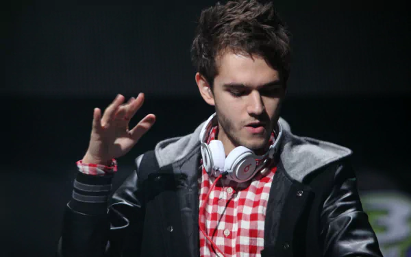 HD PC wallpaper of a music-themed DJ performing on stage: young man in a red checkered shirt and headphones around his neck, raising a hand against a dark background.