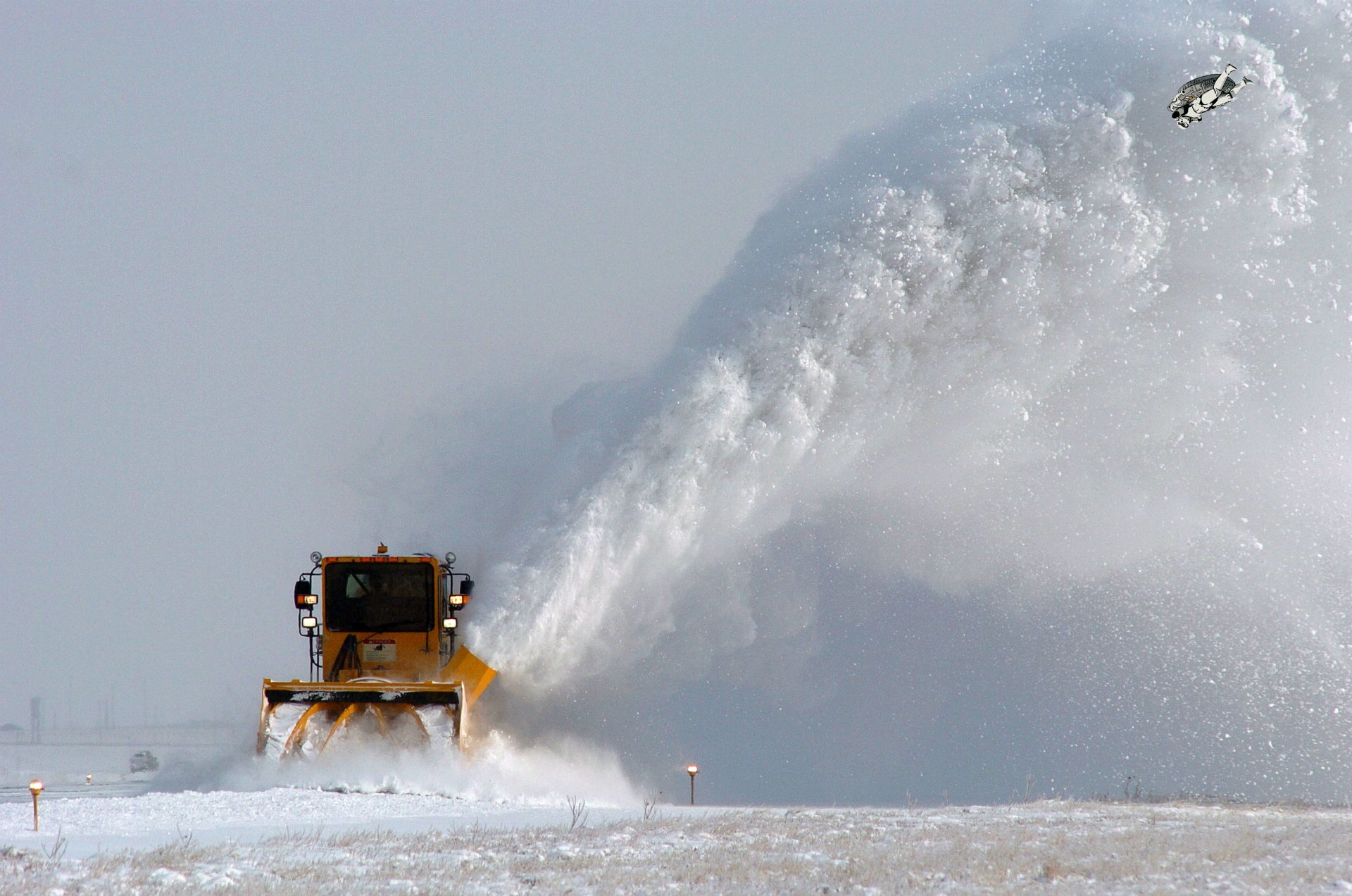 4K Ultra HD PC desktop wallpaper: yellow snowplow vehicle with front plow clearing deep snow, sending a high arcing plume against a pale winter sky.