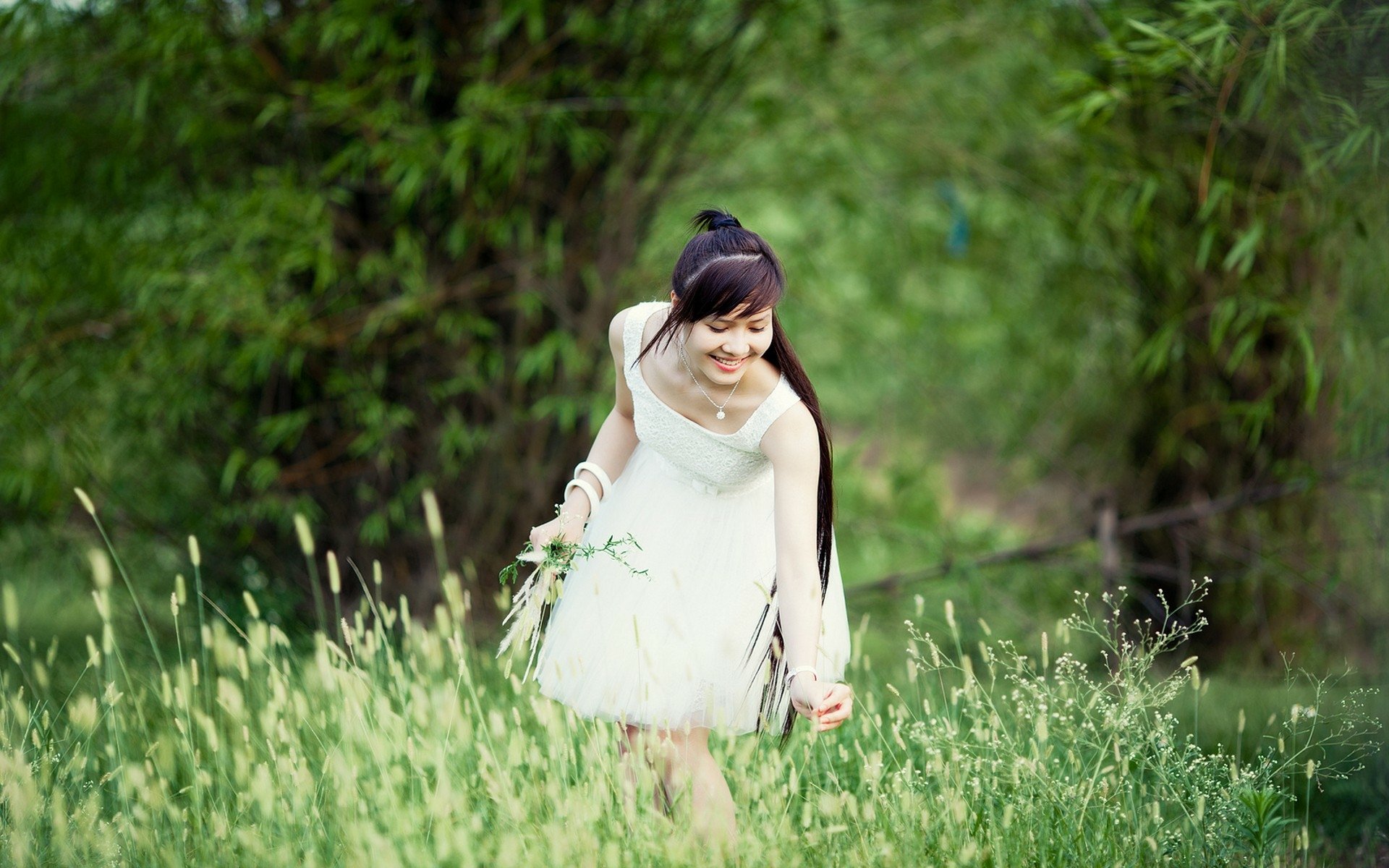 HD desktop wallpaper featuring an Asian woman in a white dress, smiling and walking through a lush green field with trees in the background.