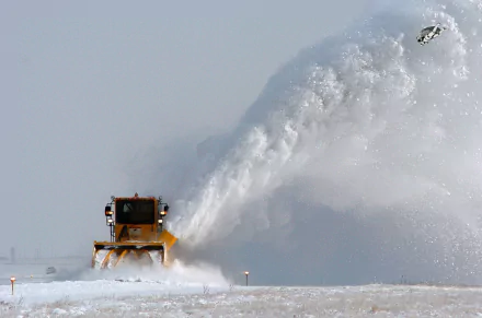 4K Ultra HD PC desktop wallpaper: yellow snowplow vehicle with front plow clearing deep snow, sending a high arcing plume against a pale winter sky.