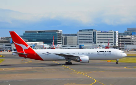 A Qantas Boeing 767 taxiing at an airport, surrounded by modern terminal buildings, capturing the essence of air travel and aviation on a vibrant HD wallpaper.