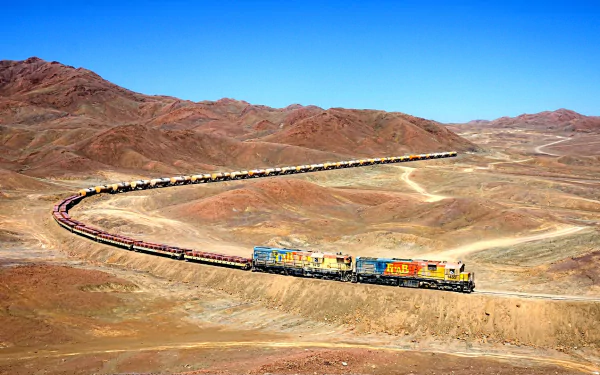 A vibrant HD wallpaper showcasing a train winding through a rugged landscape of red mountains and open terrain, highlighting the beauty of transportation in nature.