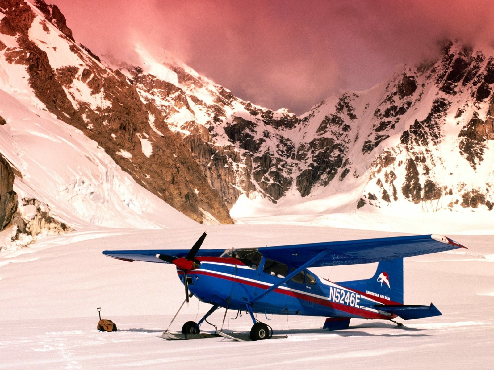 HD PC desktop wallpaper/background — blue Cessna light aircraft parked on a snowy airstrip beneath pink‑tinted alpine peaks.