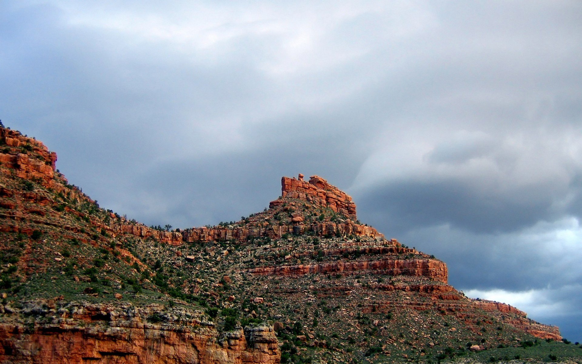 HD PC desktop wallpaper background of a red‑rock canyon mesa in a nature scene under dramatic clouds, with layered cliffs and sparse desert vegetation.