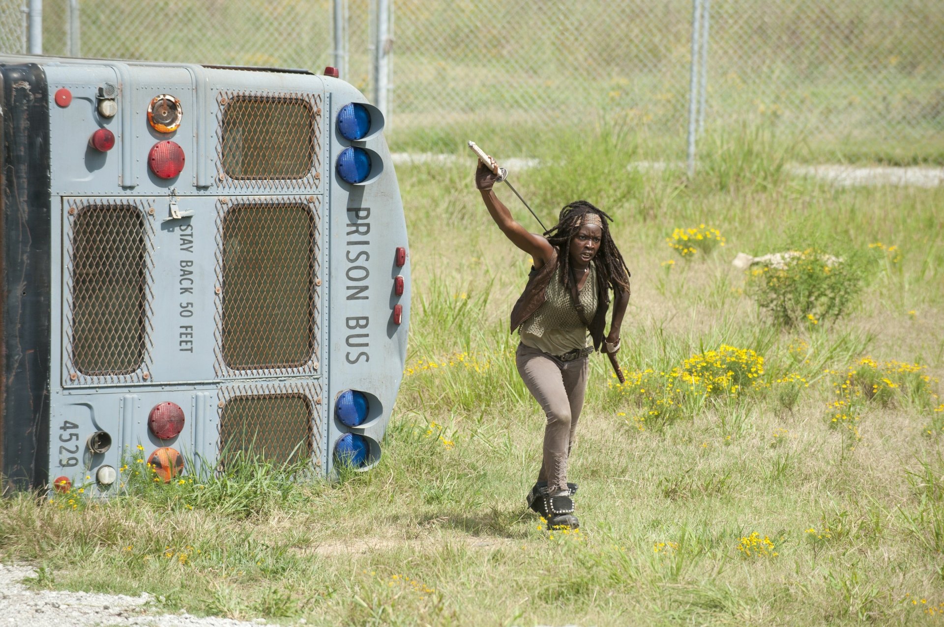 Michonne from The Walking Dead, played by Danai Gurira, runs brandishing a knife past an overturned prison bus in a grassy field in this HD PC desktop wallpaper.