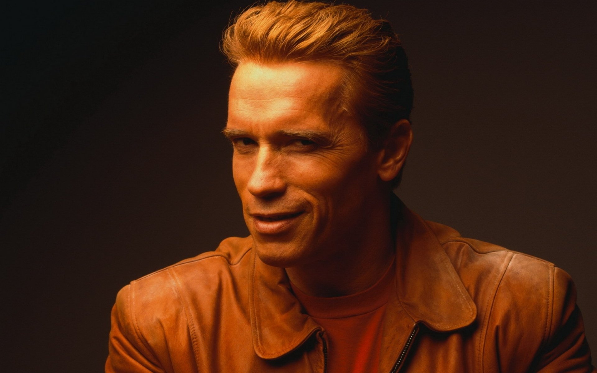 HD PC desktop wallpaper featuring celebrity Arnold Schwarzenegger with slicked-back hair and a confident expression, wearing a brown leather jacket against a dark background.