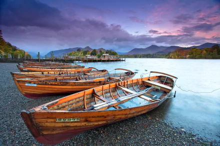 HD PC desktop wallpaper showing wooden boats moored on a calm lakeshore with mountains and a colorful sunset sky in the background.