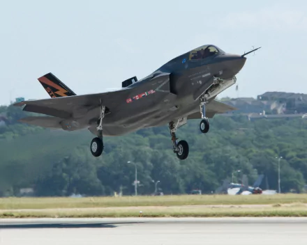 Military Lockheed Martin F-35 Lightning II fighter jet captured in mid-landing, set against a clear sky and lush green background, featured as a HD PC desktop wallpaper.