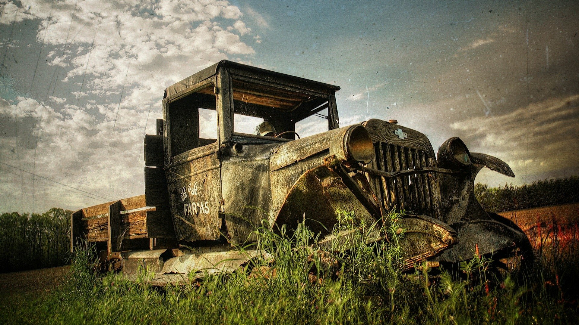 HD PC desktop wallpaper featuring an old, rusted vehicle wreck resting in a grassy field under a cloudy sky.