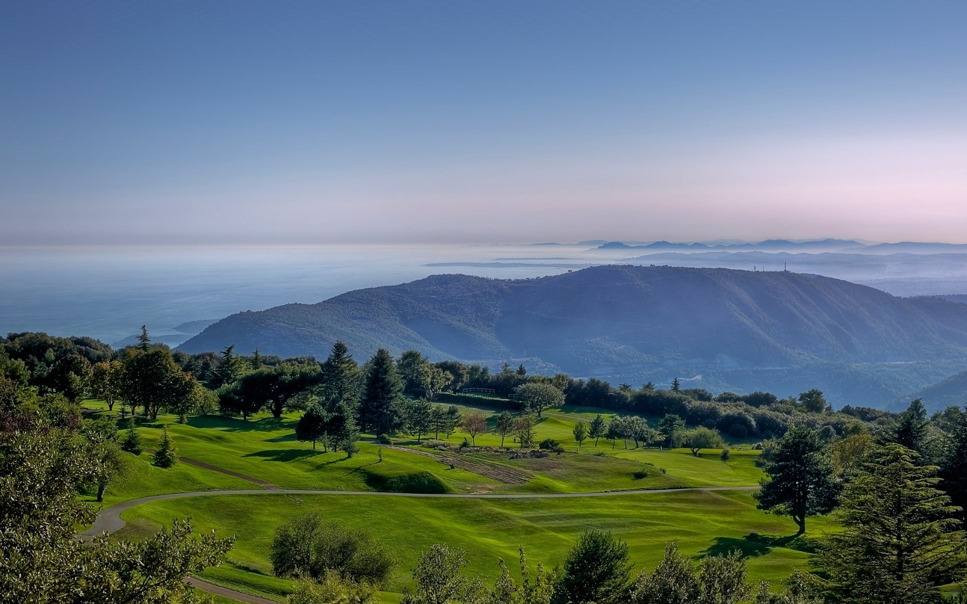 2K Quad HD PC desktop wallpaper — nature landscape of rolling green hills and scattered trees descending to misty blue mountains under a wide, clear sky.